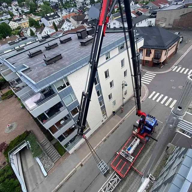 Lang kran løfter utstyr fra lastebil i bygate, sett ovenfra med bybebyggelse rundt.