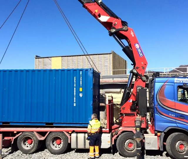 Blå lastebil med kran løfter en blå container på byggeplass med fjell og sol i bakgrunnen.