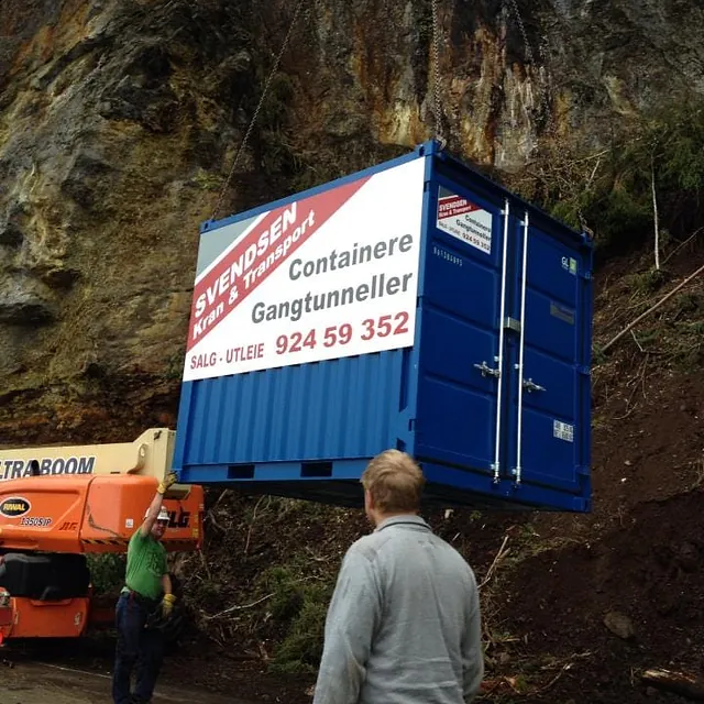 Blå container med reklameskilt heises opp foran fjellvegg, to personer står og følger med.