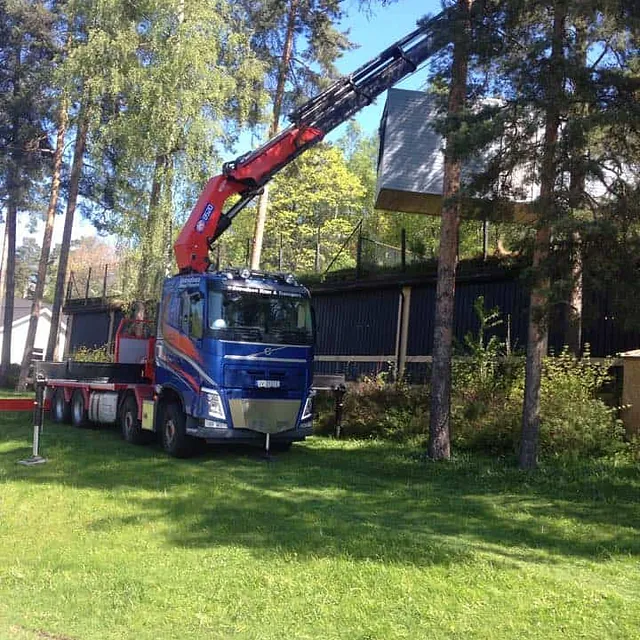 Kranbil står på grønt gress mens kranarmen strekker seg over trær mot bygning.