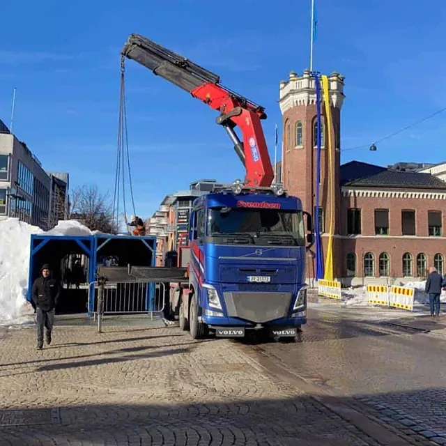 Kranbil plasserer blå gangtunnel ved gate i byområde, omgitt av snø og bygninger.