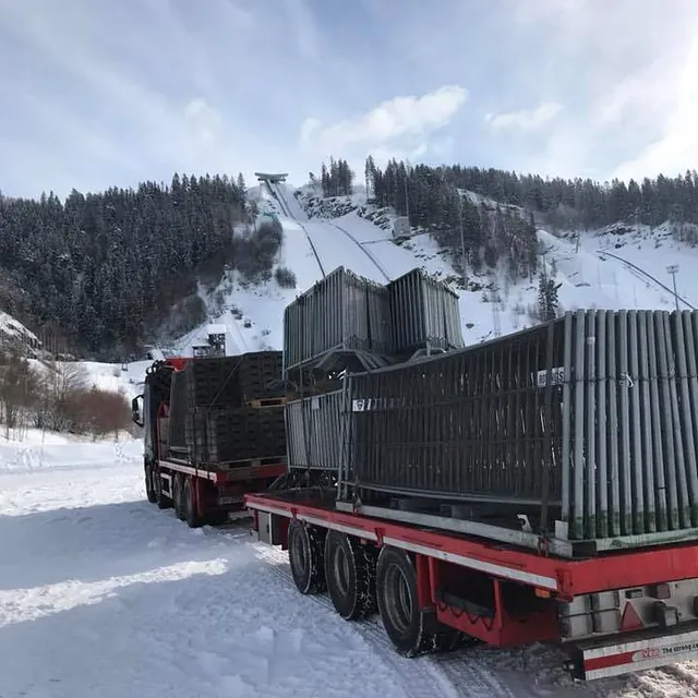 Lastebil med tilhenger fullastet med stålgjerder parkert i snødekt terreng ved skihoppbakke.