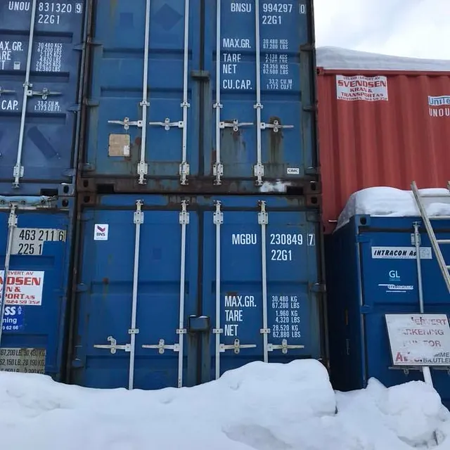 Flere stablede fraktcontainere i blått og rødt, omgitt av snø og skilt med tekniske spesifikasjoner.