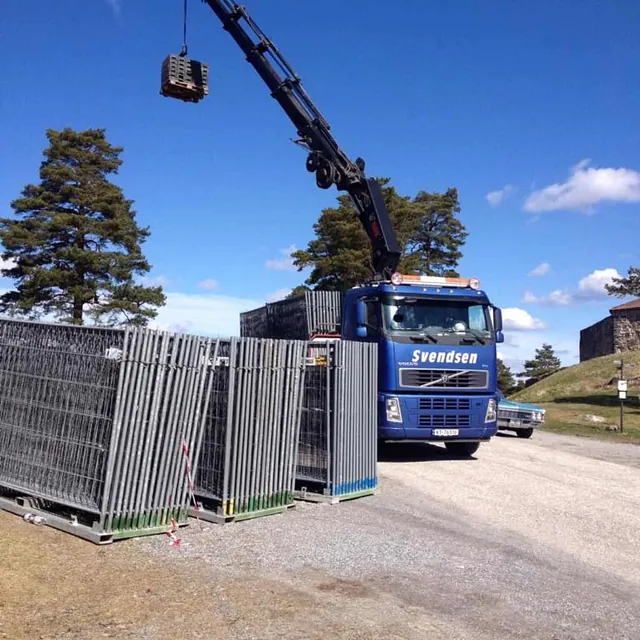 Lastebil med kran løfter en pall i lufta, gjerdeelementer står stablet ved siden av.