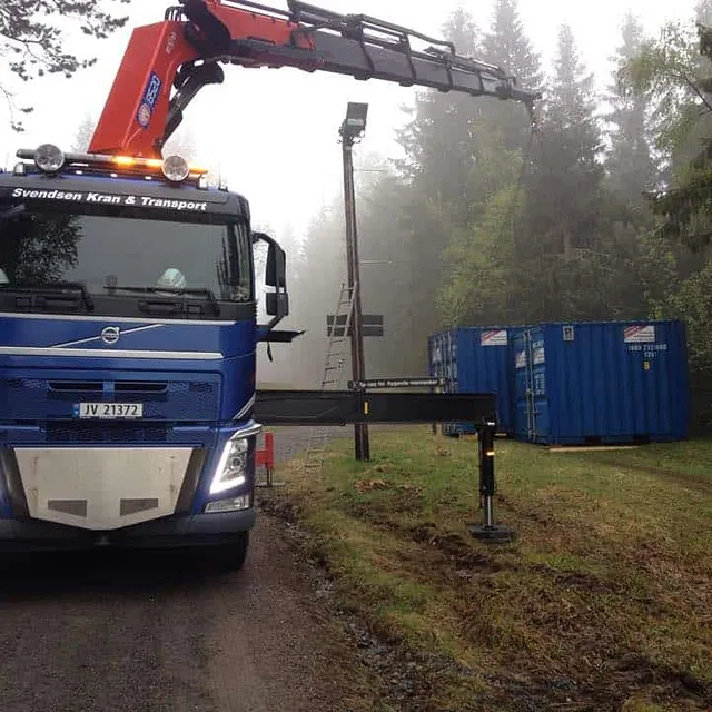 Lastebil med kran står ved skogkant og gressplen, to blå containere i bakgrunnen.