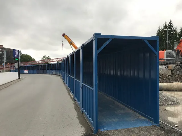 Lang blå gangpassasje av stål ved vei og byggeplass, med kran og maskiner i bakgrunnen.