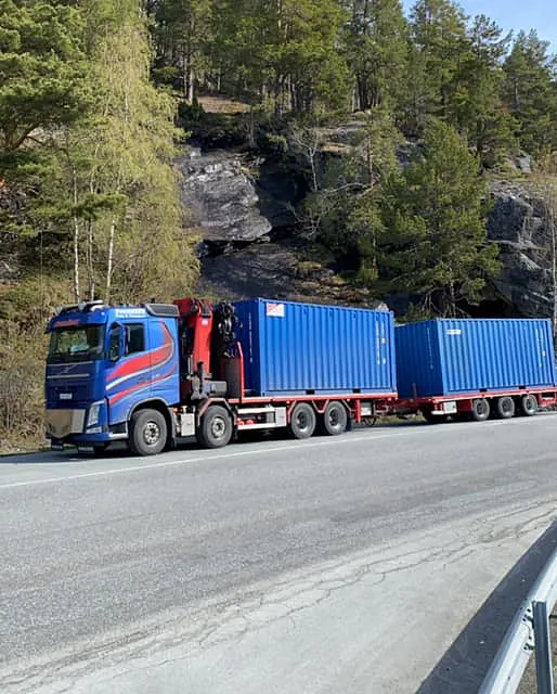 Lang lastebil med blå førerhytte og røde detaljer frakter to blå containere på landevei.