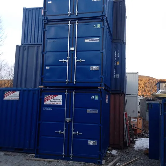 Stablede blå containere i tre høyder på gruslagt område, omgitt av andre containere og utstyr.