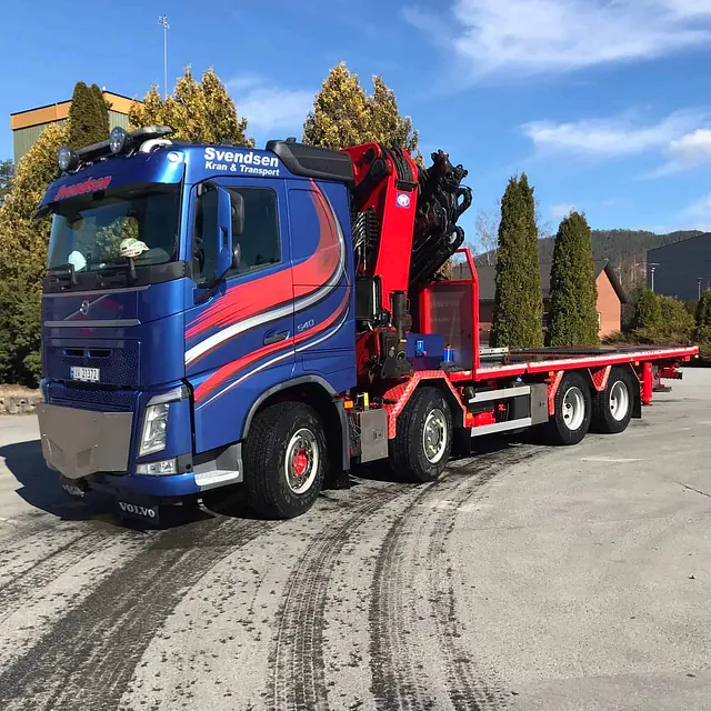 Svendsen kranbil med flat henger og løftekran parkert på asfalt i solskinn.